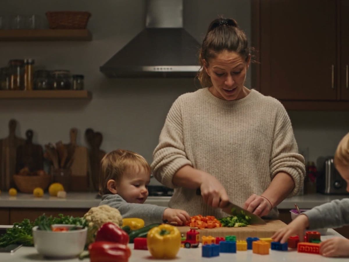 A parent chopping vegetables at the kitchen counter while a toddler plays with lego beside the cutting board